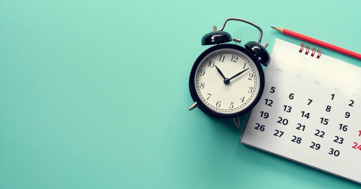 Closeup of an old-fashioned alarm clock next to a calendar and a pencil lying on a seafoam green background.