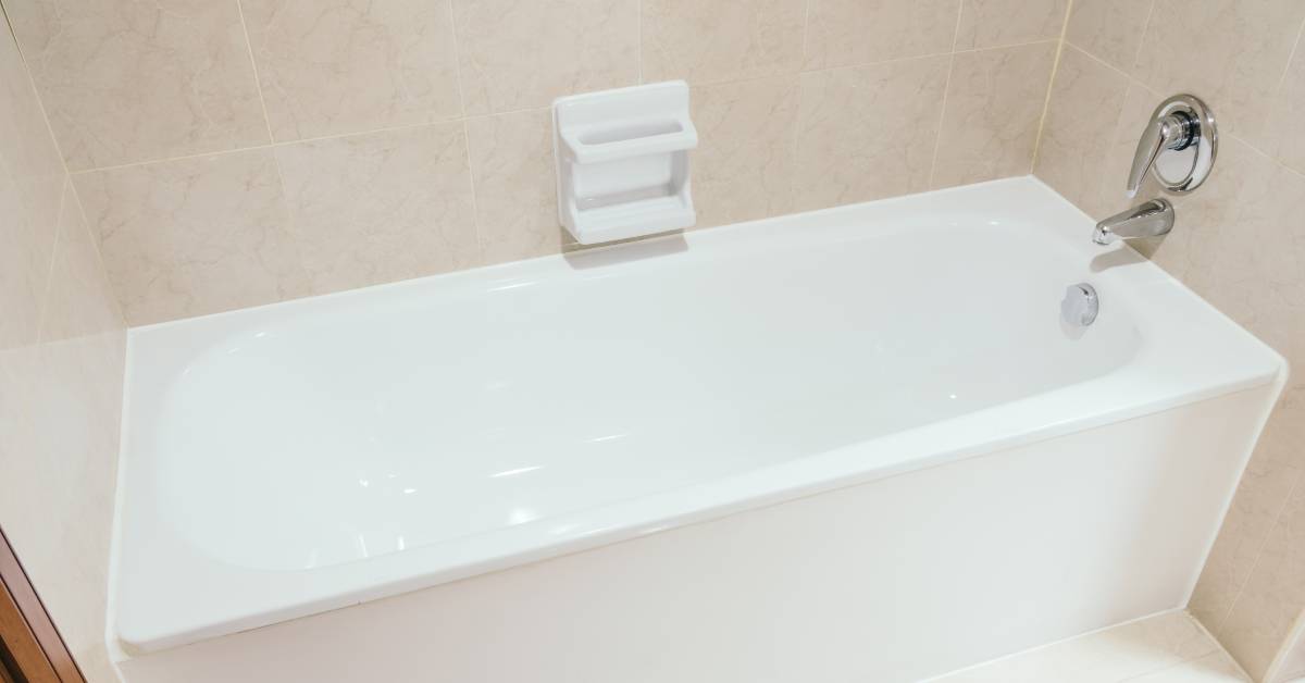 Closeup of a white ceramic bathtub with chrome hardware, large tan tiles, and a white soap dish on the wall.
