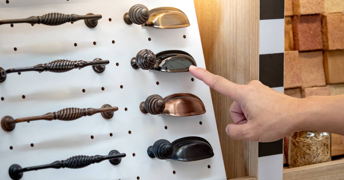 Closeup of someone’s hand pointing at a vintage-style cabinet handle that is next to various other cabinetry hardware options.
