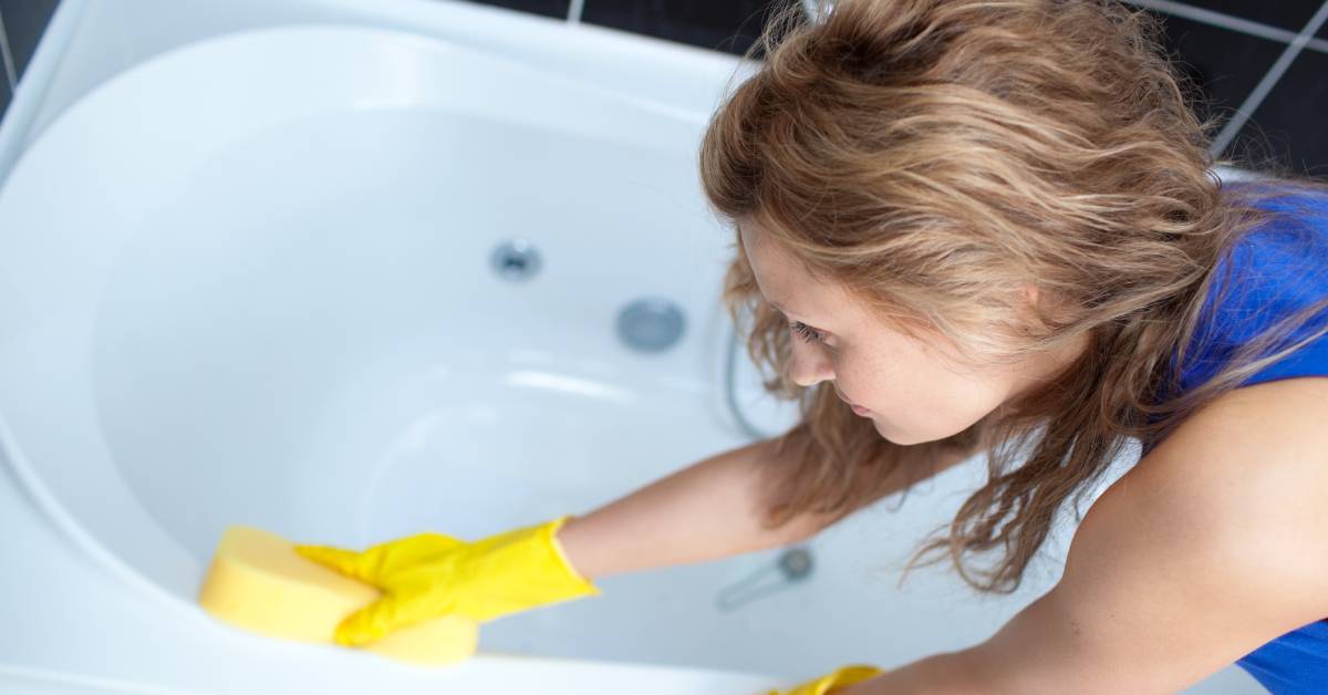 Closeup of a woman with auburn hair and a blue shirt wearing yellow rubber clothes while cleaning a bathtub.