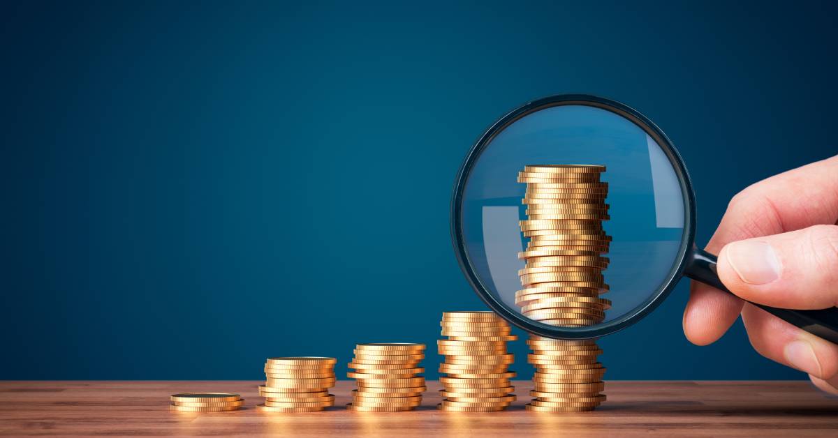 A close-up of five tiered stacks of coins sitting on a table and someone holding a magnifying glass looking at the coins.