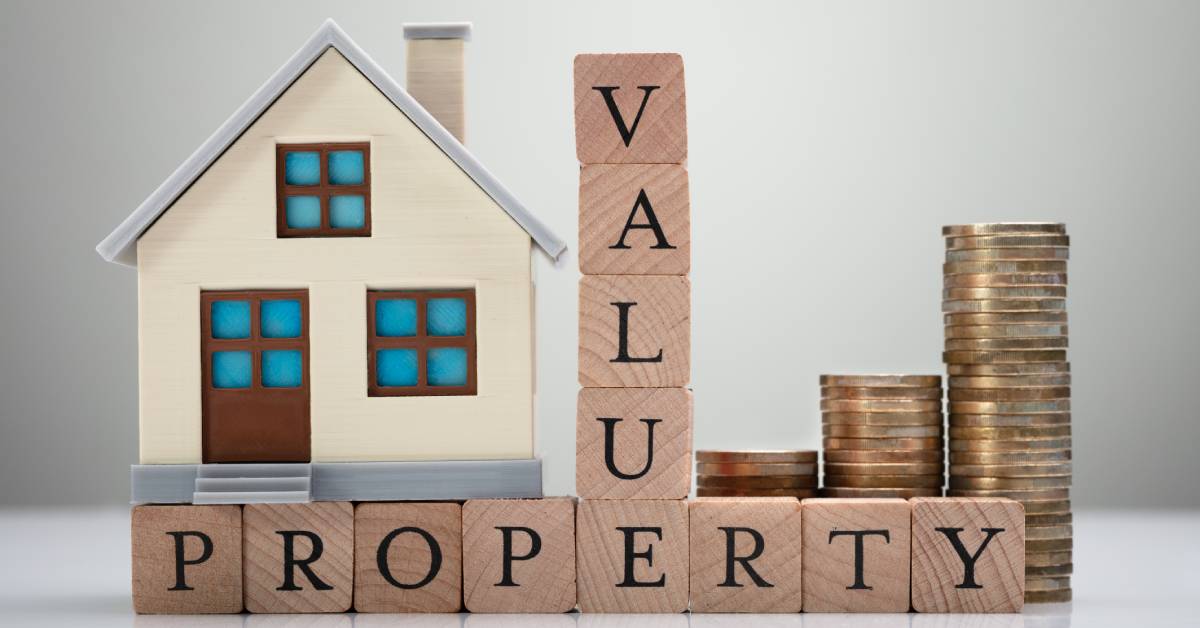 Close-up of several blocks stacked together that say “property value” with a small house and stacks of coins behind them.