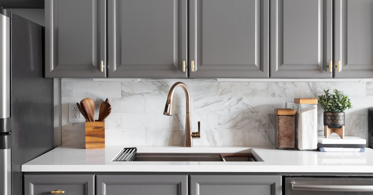 Closeup of a modern kitchen sink, white countertops, and grey cabinets with elegant gold hardware pulls.
