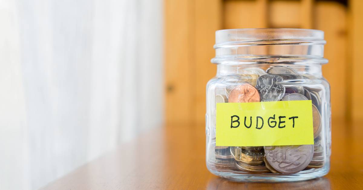 Closeup of a clear mason jar filled with coins and a yellow label on the front that says the word “budget” in black text.
