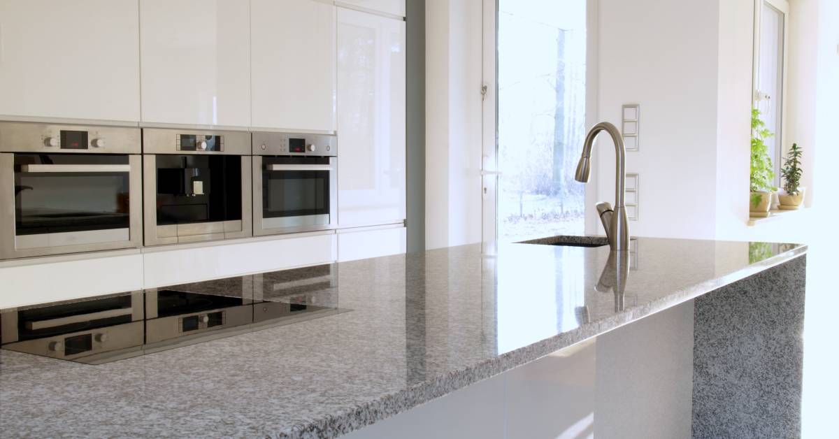 Close-up of a large, modern kitchen island with granite countertops and three ovens in the background.
