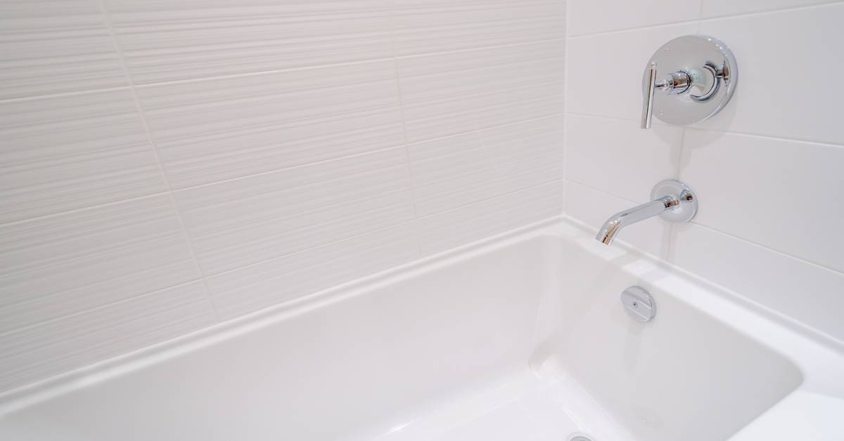 Close-up of a modern, clean, white bathtub with white textured wall tiles and a chrome faucet and finishes.