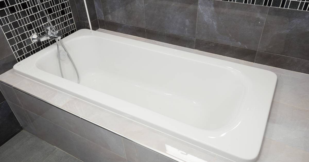 Closeup of a clean, white ceramic bathtub with chrome hardware surrounded by grey and black tiles that go up the wall.