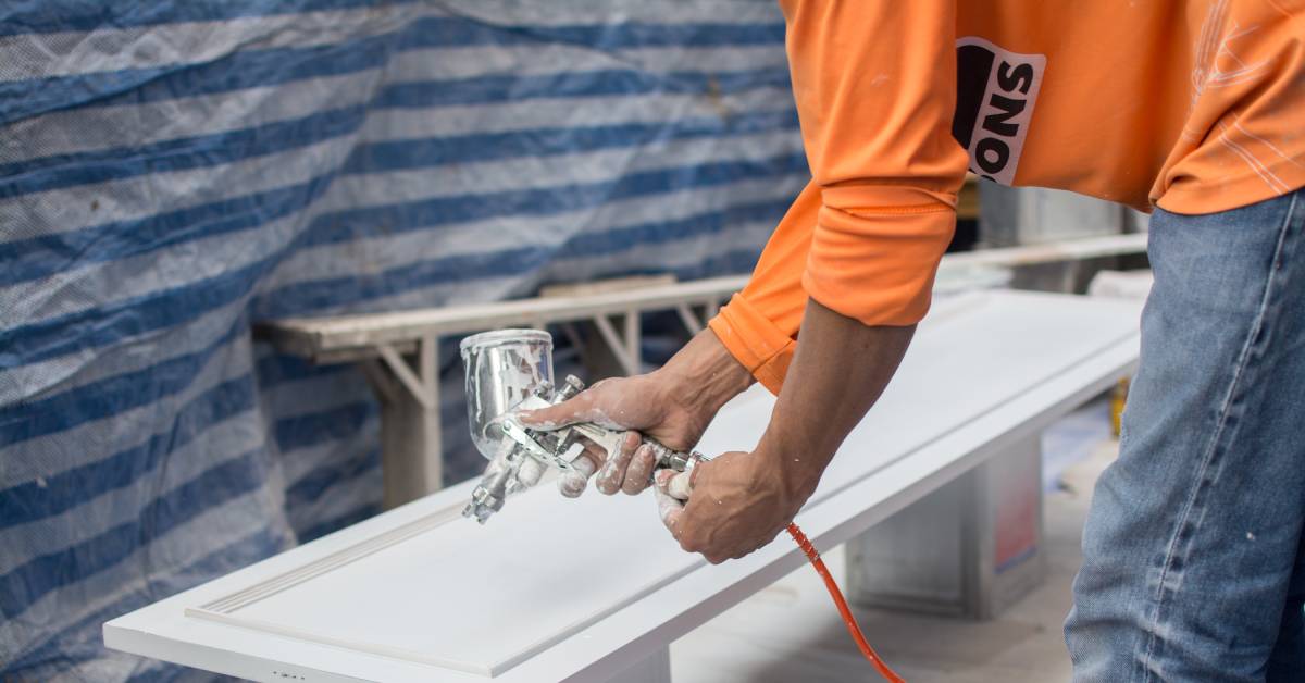Closeup of a man in jeans and an orange shirt using a spray gun to repaint cabinets in a white color.